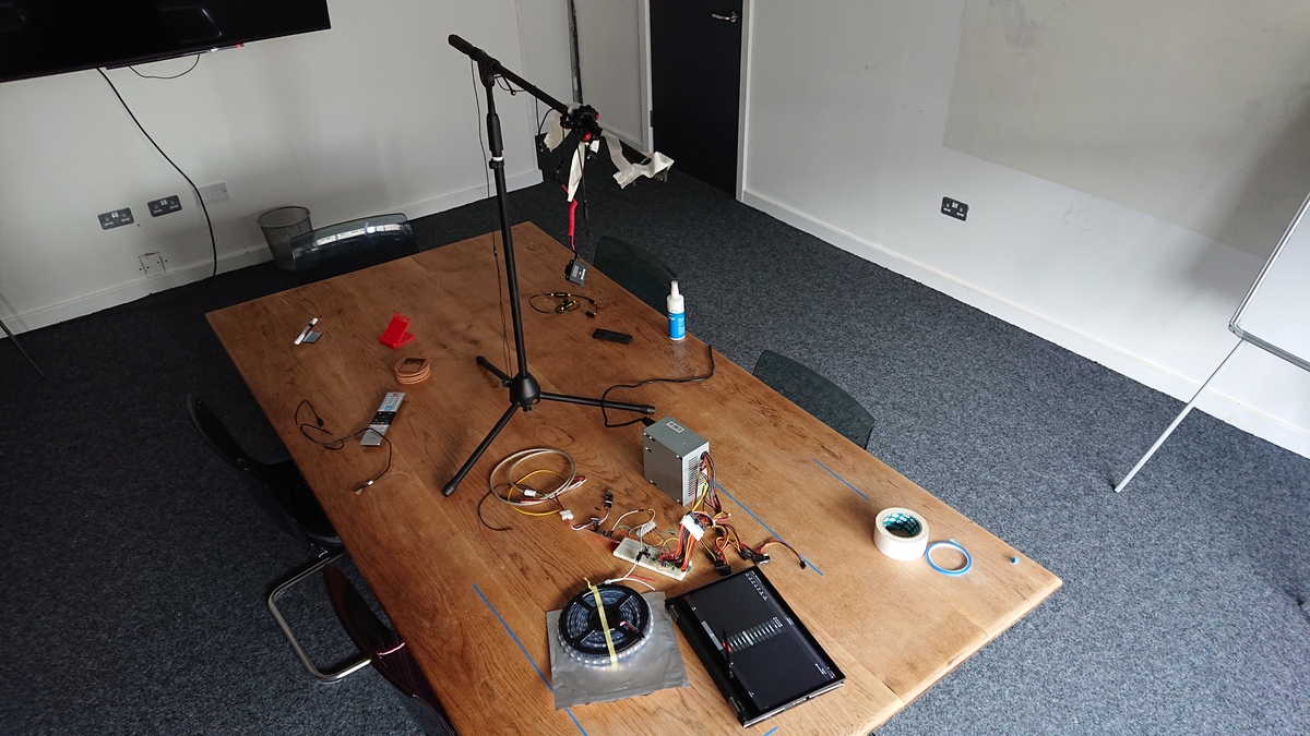 Photo of a room with a big wooden table in the centre, littered with LEDs, a prototype circuit, power supply and a laptop, with a microphone stand repurposed as a camera tripod
