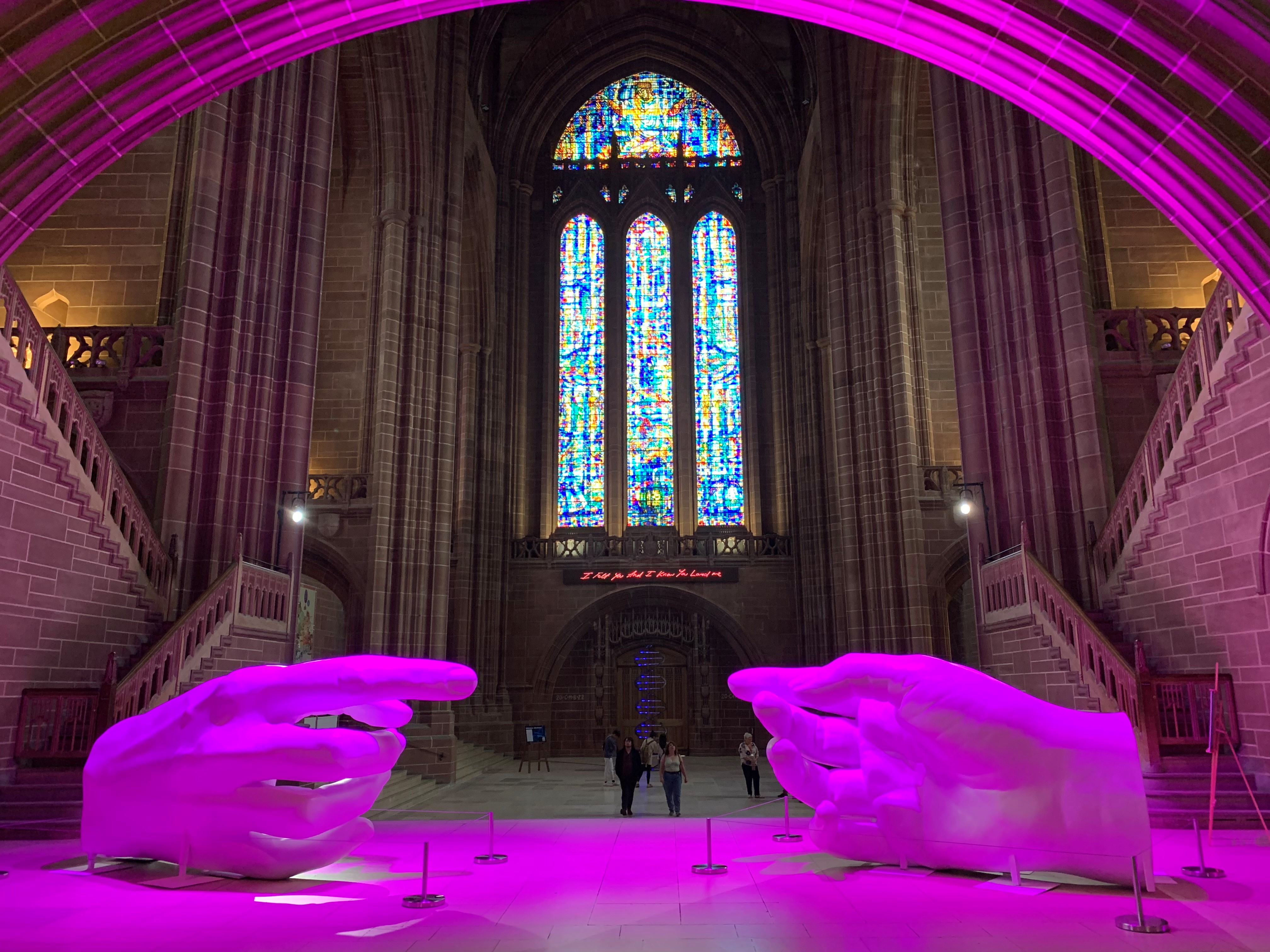 The inside of a large sandston cathedral.  A sculpture is centre-stage of two huge female hands with their forefingers reaching towards each other to make contact.  The scene is lit with a wash of purple light.