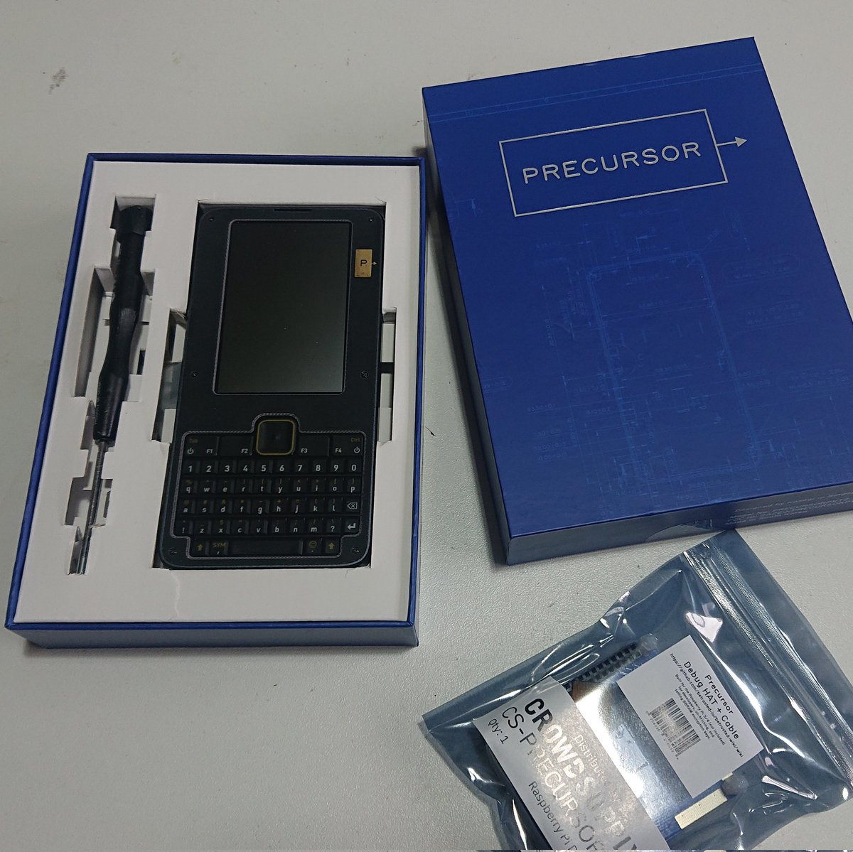 A box labelled 'Precursor' is open on a table revealing a device with a screen and qwerty keyboard that looks like a Blackberry phone. Next to it is a small screwdriver. Below the box is a small circuit board in an anti-static bag