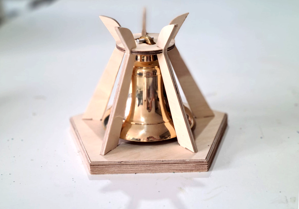 Brass bell on a pentagonal plywood base supported by five shaped upright supports.