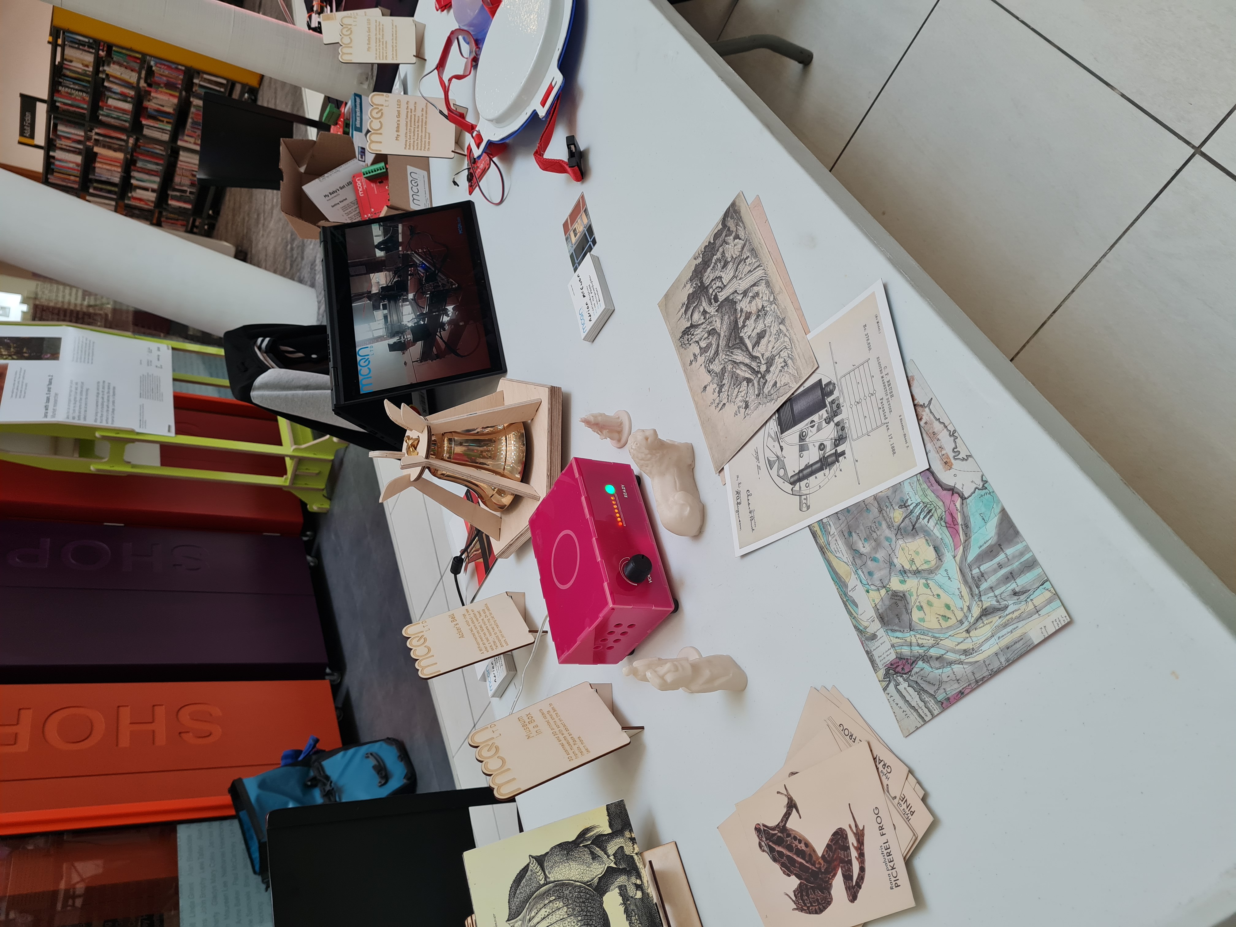 A table featuring several MCQN projects including Museum in a Box - a pink box with a volume dial, surrounded by post cards and 3D printed objects which can be used with it and the Ackers Bell - a brass bell in a pentagonal housing. There is also a screen showing video and the edge of the Peleton MBGL disco breastplates can be seen on the edge of the frame.