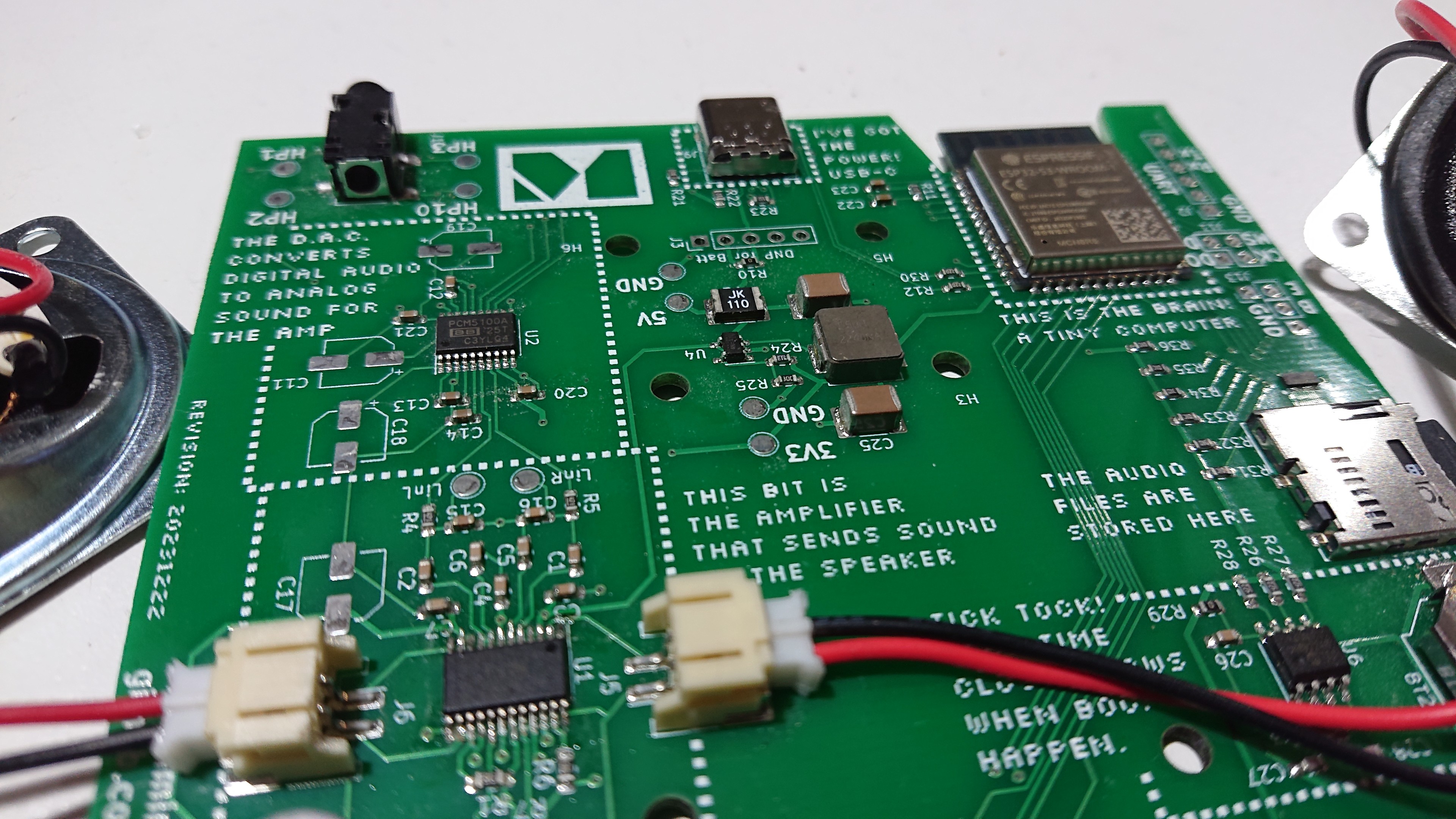 A green printed-circuit board populated with an assortment of chips and other components.  The text on the silkscreen says things like 'This bit is the amplifier that sends sound to the speaker' and 'This is the brain!  A tiny computer'