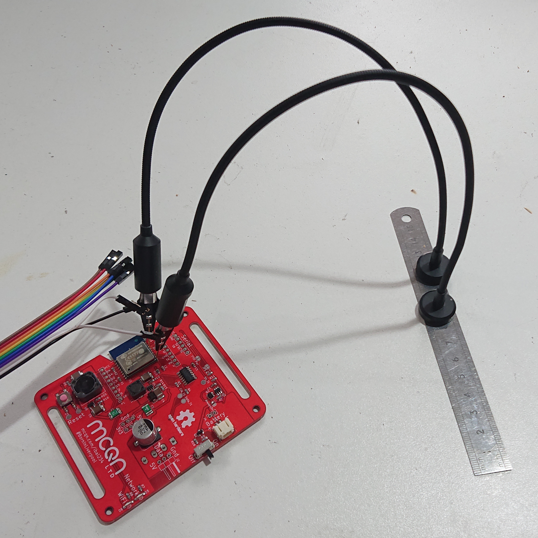 A red PCB on a white table with two probes placed onto points on the PCB.  The probes are held in place with gooseneck connectors which arch over to magnetic bases which are held steady on a steel ruler
