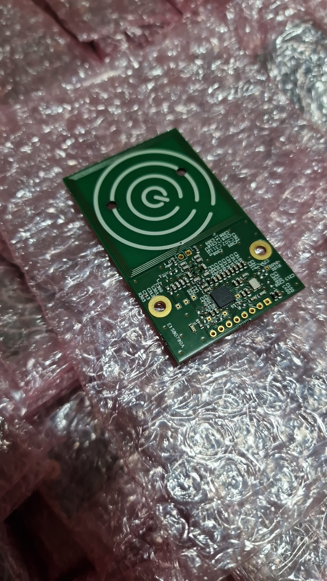 A rectangular green PCB resting on a sheet of pink bubblewrap.  Just over half the board is taken up with a set of concentric white circles of silkscreen, with a Q in the centre.  The remaining board area has assorted electronic components packed into it