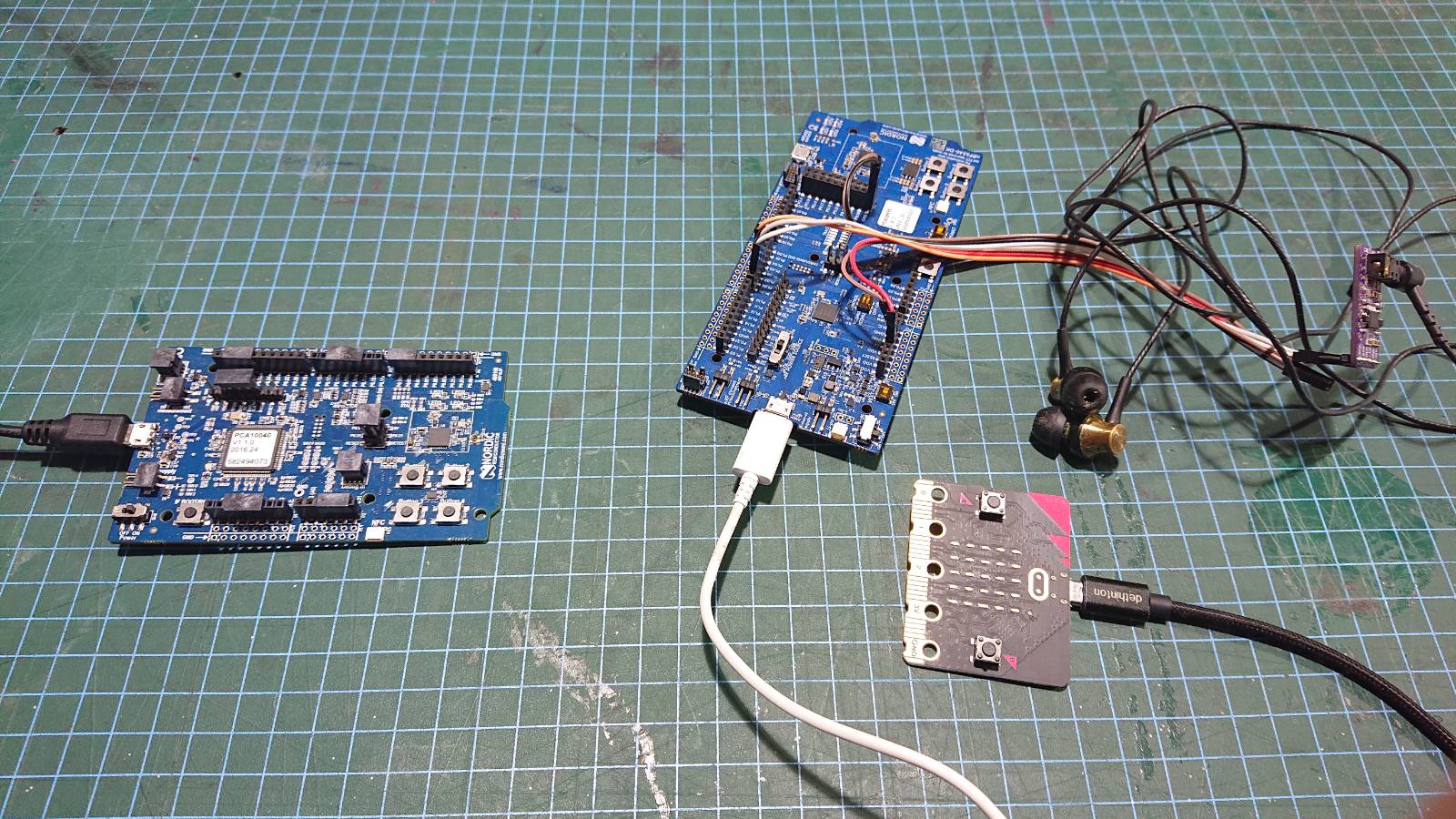 A few different nRF dev boards laid out on a cutting mat.  On the left is an nRF52dk, which is broadcasting the audio over Bluetooth.  On the right are two other boards which are both receiving the broadcast: an nRF5340dk, which is wired to an I2S DAC board with headphones plugged in; and a micro:bit v2, which will—in time—show the audio levels on its LED display