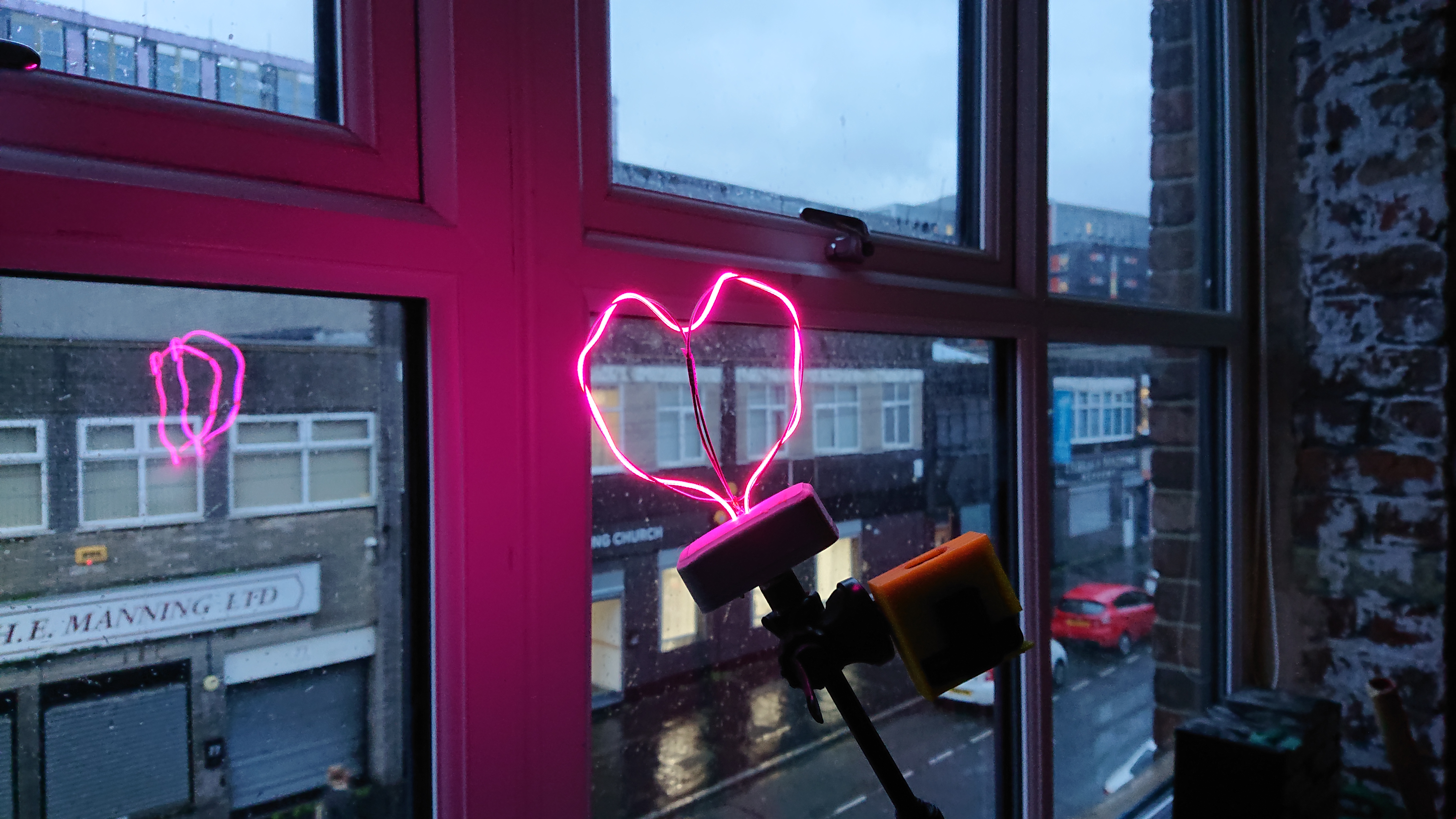 We're in a dimly-lit room looking through the window onto a grey rainy street as the light fades.  Being held in front of the window is a selfie stick with a wireless transmitter clamped to the top of it and the Heartlight screwed on the end and lit up in shocking pink