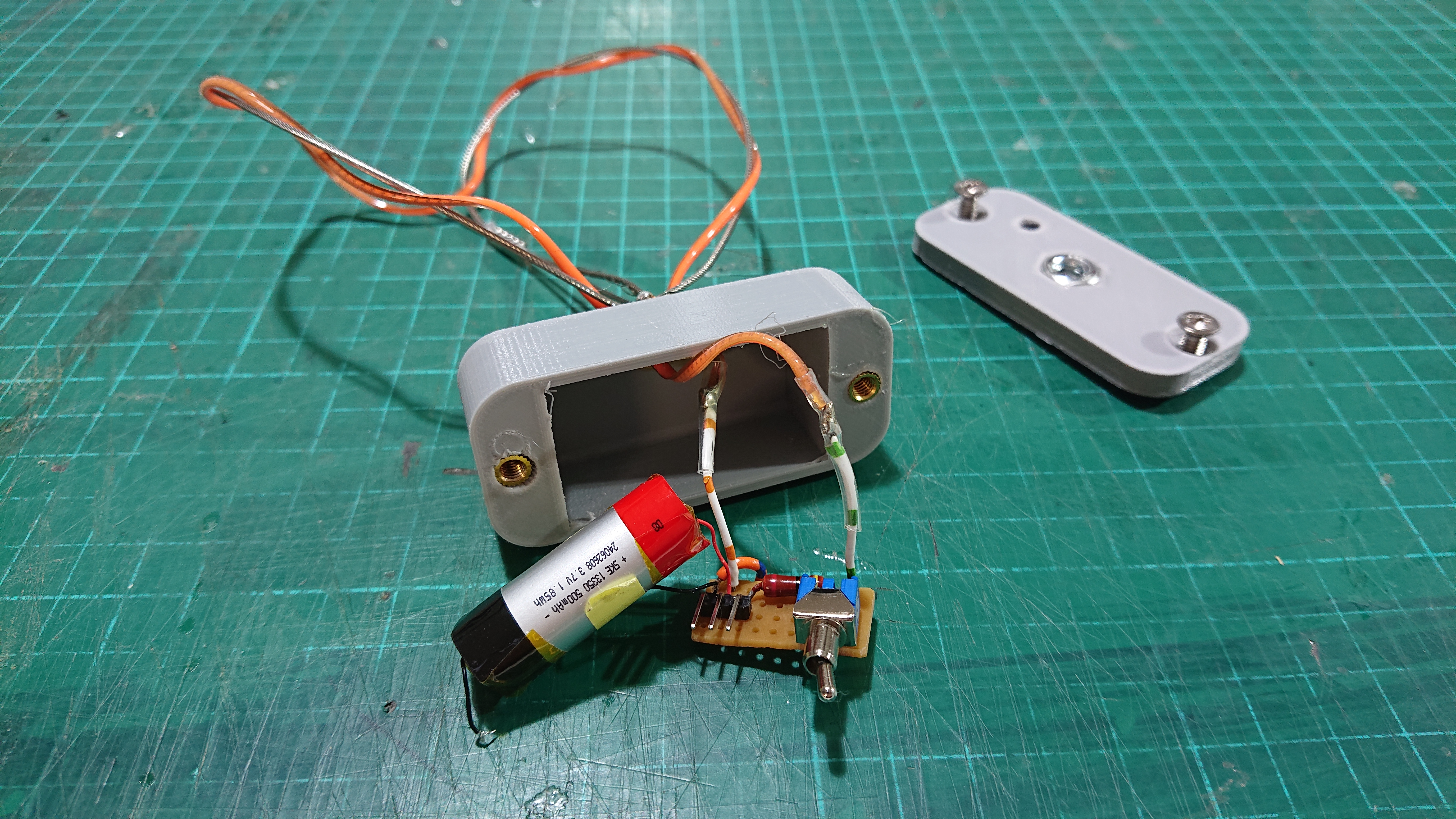 The partially disassembled Heartlight sits on a cutting mat.  The base is 3D printed in grey plastic and is open at the moment, with a small cylindrical battery, resistor, charging pins and toggle switch pulled out so they're more visible.  The components are connected on a small piece of veroboard and there are two wires coming off that which feed through the plastic base to the LED noodle that's wrapped round a wire frame on the other side