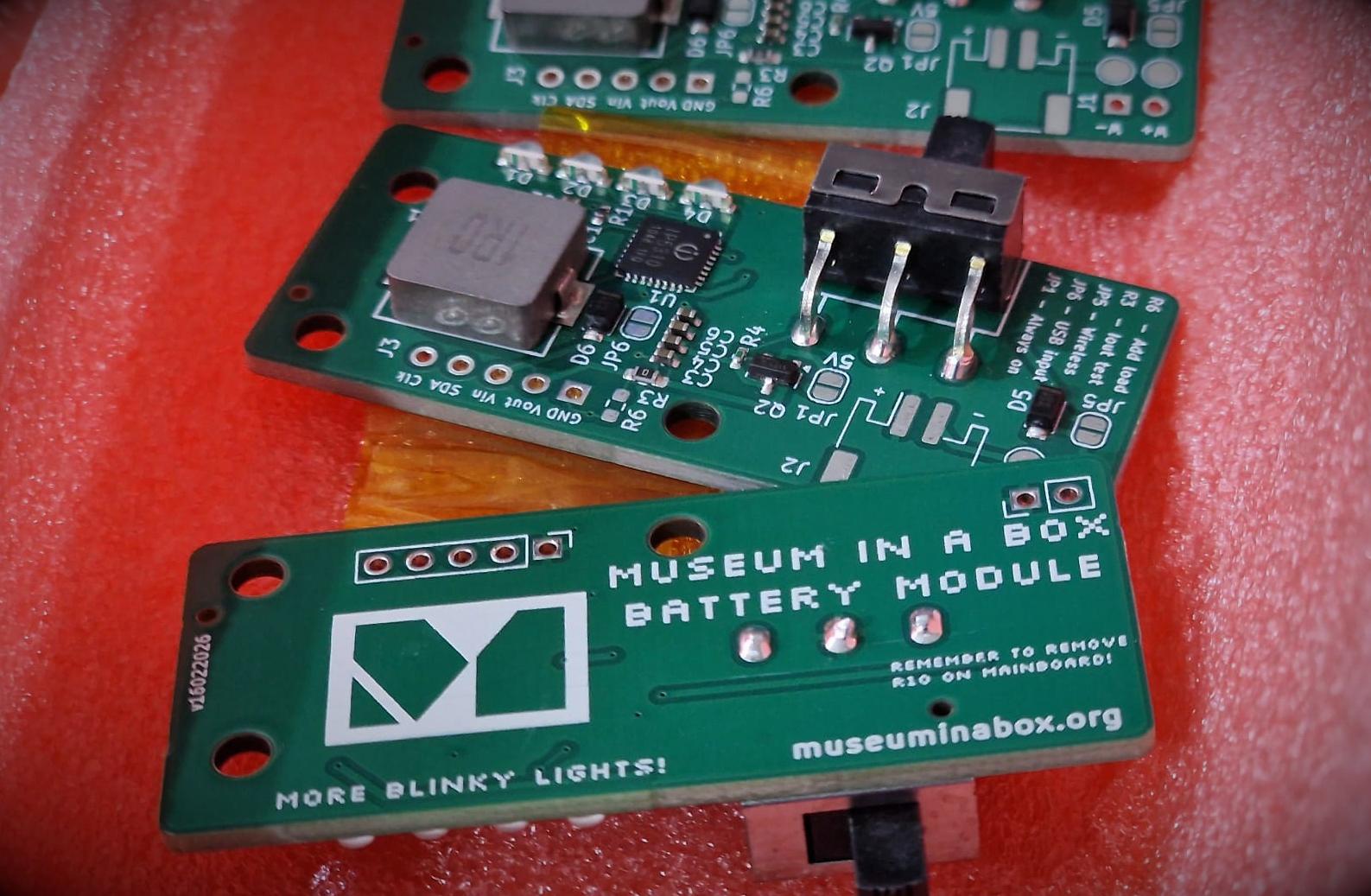 Three, of five, green PCBs that allow for battery power. The foreground shows the front with a Museum in a Box logo and the text 'Museum in a box battery module' and 'More blinky lights' The other boards show the reverse with a chunky on off switch, some LEDs and a battery power management IC.