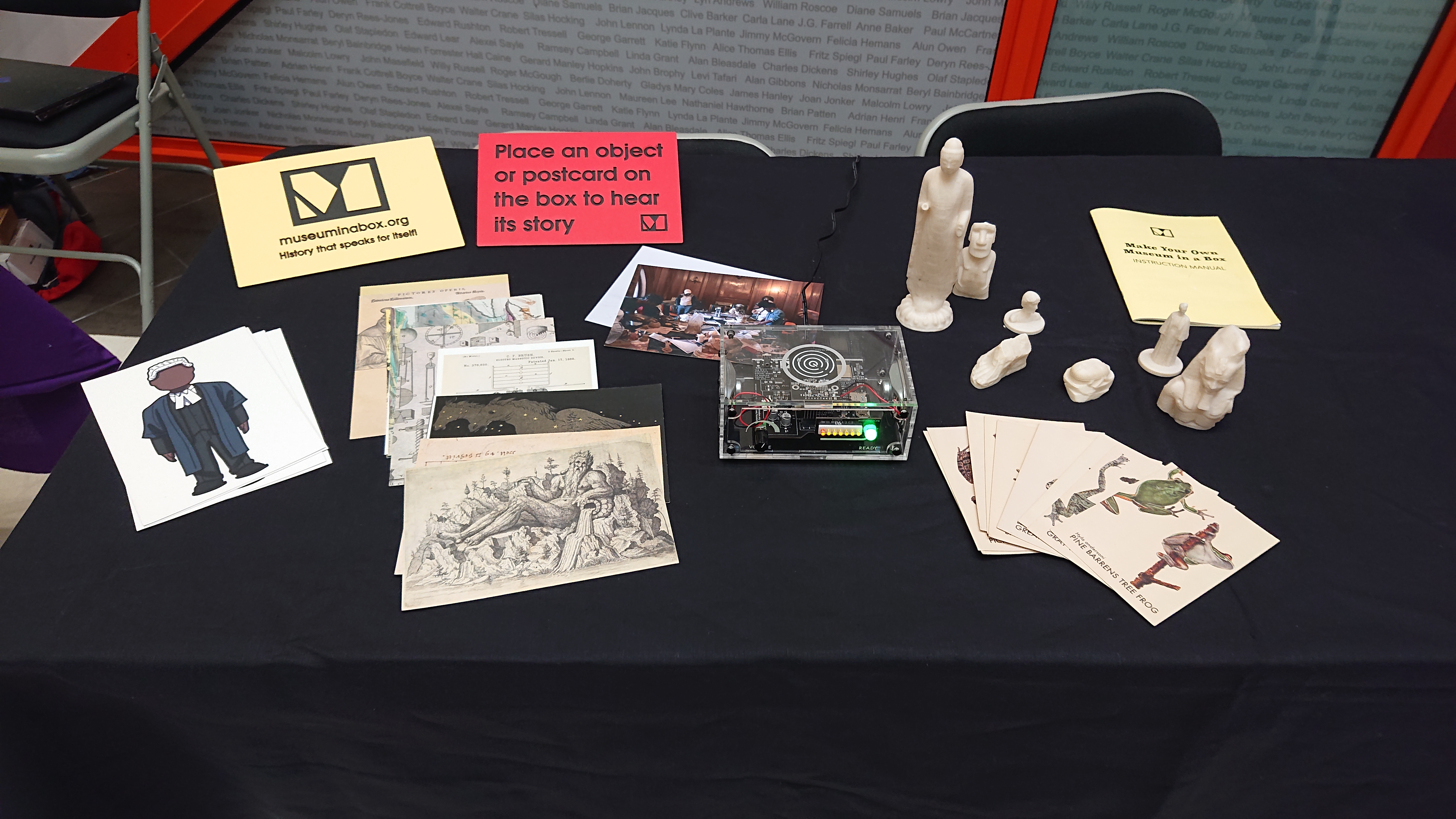 A transparent Museum in a Box is sat on a table surrounded by an array of 3D prints of statues and postcards of frogs, a barrister, reproduction artworks and a photo of some people around a table of artefacts.  At the back of the display are two signs: one with the Museum in a Box logo and URL, and one reading 'Place an object or postcard on the box to hear its story'