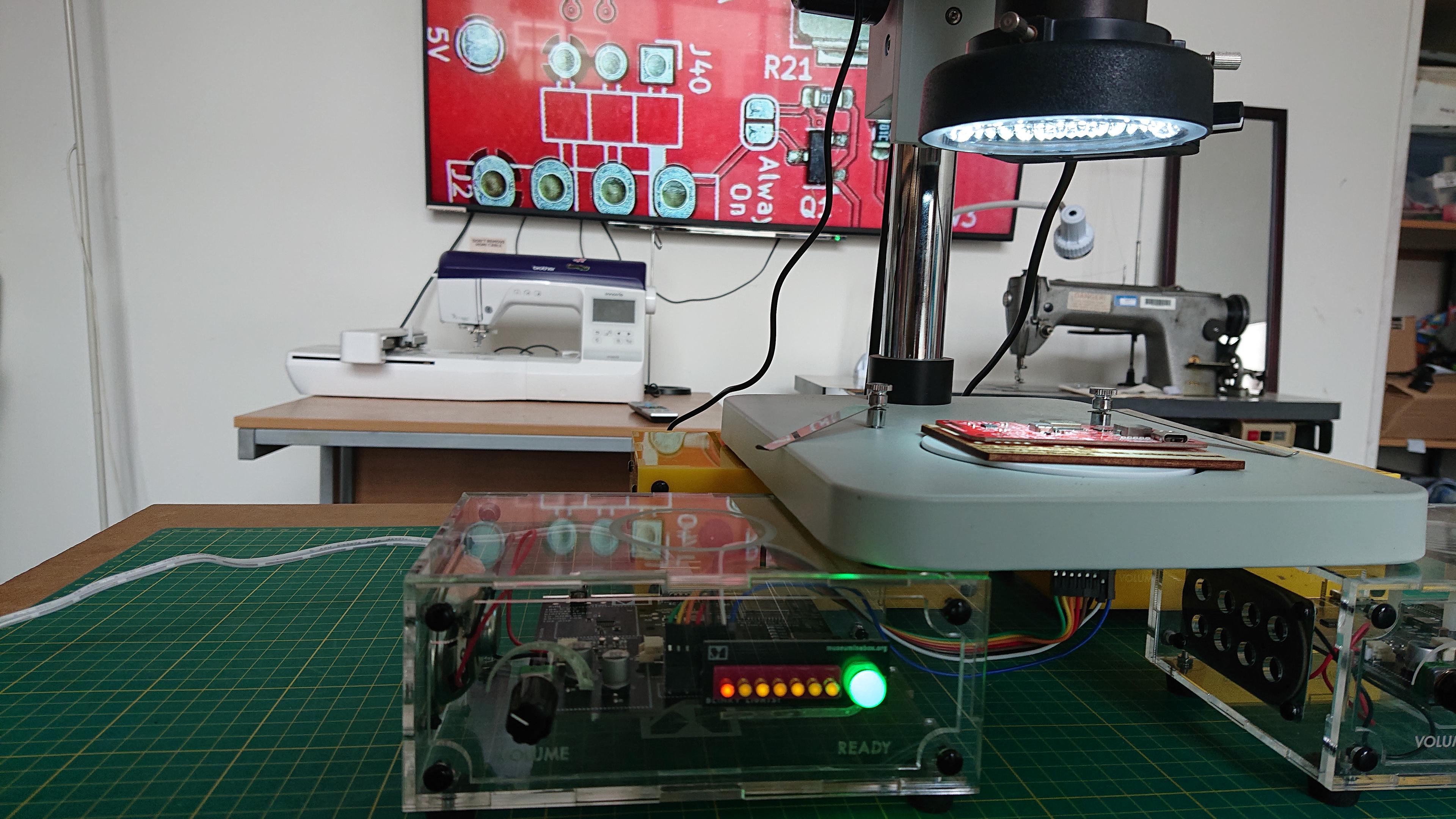 A microscope (we can only see the lower half of it) is sat upon a quartet of Museum in a Box boxes (look, we have them readily to hand).  The Museum in a Box in centre of the frame is turned on (the LEDs on the front of it are all lit up) and some wires sneak out from the circuit board inside it to a position just under the stage of the microscope.  There's a red circuit board (one of our My Bike's Got LED boards, as it happens) sat on the microscope stage.  In the background there's a massive telly on the wall, and it's showing the output from the microscope: a close up of the red PCB.  There's also a CNC embroidery machine and an industrial sewing machine in shot, but they're not important right now.