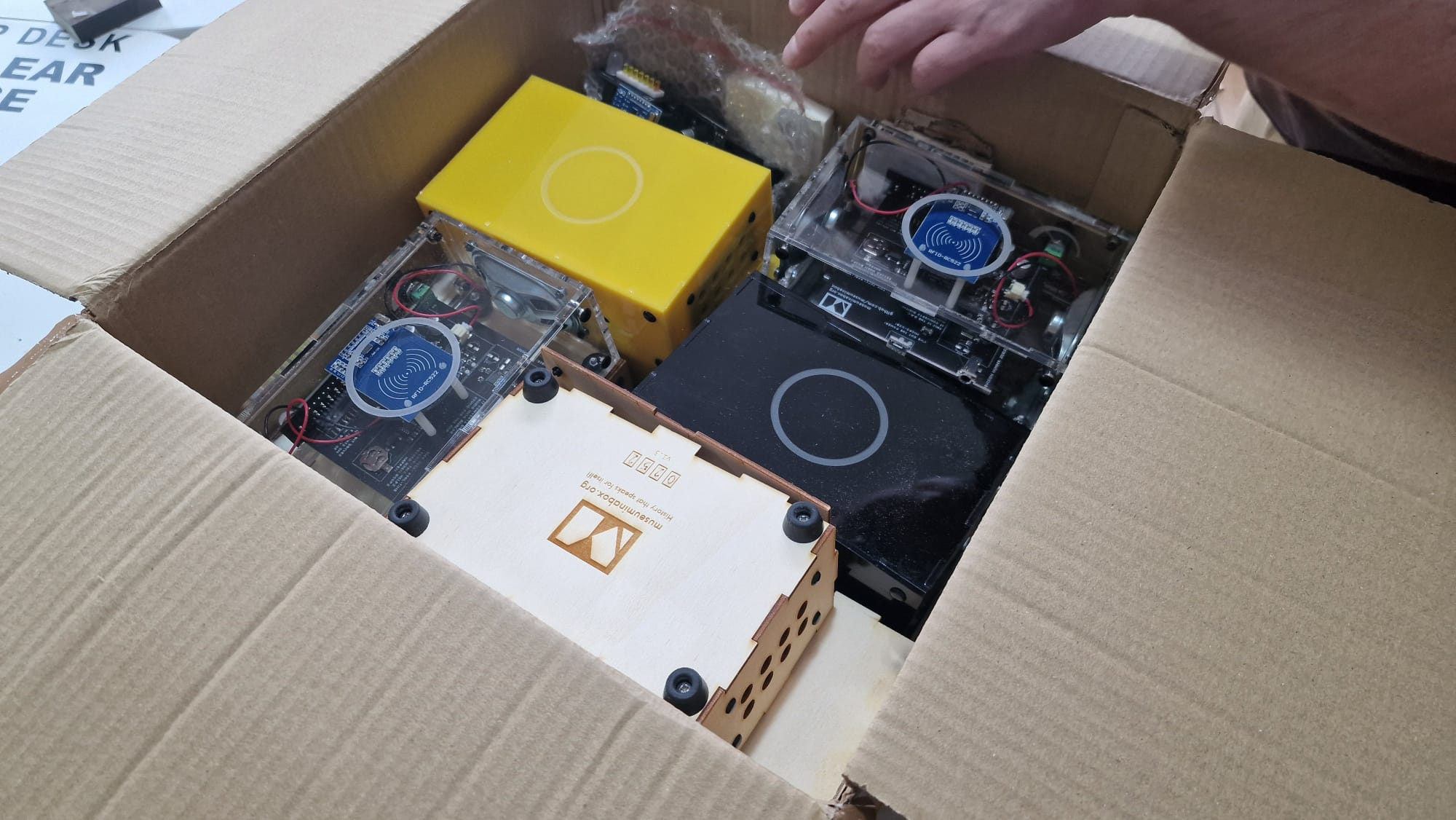 An open cardboard box showing a collection of Museum in a Box boxes inside: a couple of transparent ones, a yellow one, a black one and a wooden one. The front of the wooden one can just be seen to not be connected to the rest of the box, and in a corner there's a seemingly flat-packed version reduced to its component parts
