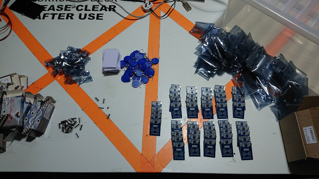 A workbench covered with an assortment of parts and packaging, grouped into clusters: empty boxes; anti-static bags; white RFID 'credit card' tags; blue 'key fob' RFID tags; and soldered RFID readers, arranged in rows of five