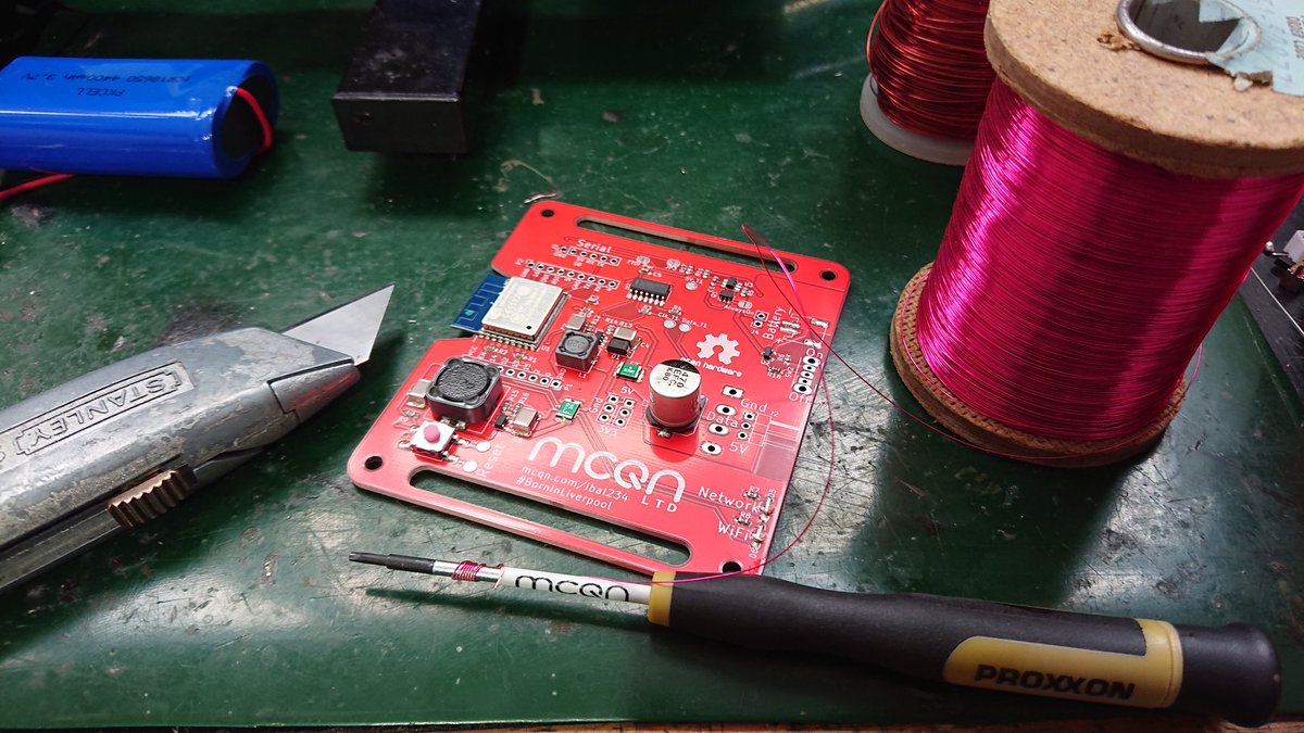 A spool of shiny pink enamelled wire with some wrapped round the shaft of a screwdriver. Next to that lies the Stanley knife which has scraped off some of the enamel. In the middle is a red PCB waiting to be fixed