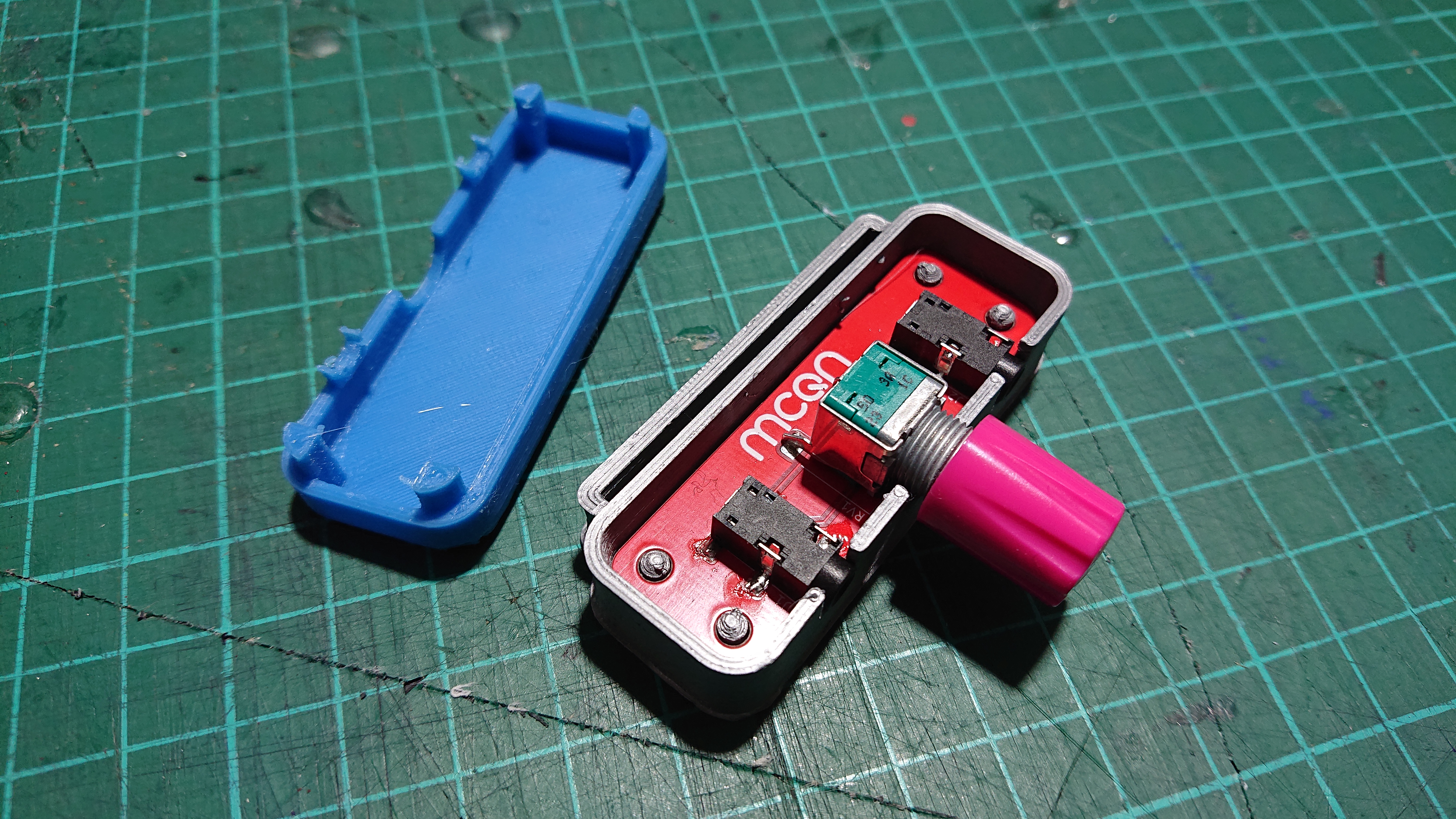 The two-tone case from the last image is shown with the blue top half removed and placed on the table upside-down.  That shows a red PCB with the MCQN logo on it inside the silver bottom half, and the audio jack sockets and stereo potentiometer visible on the PCB, poking through the side of the case.