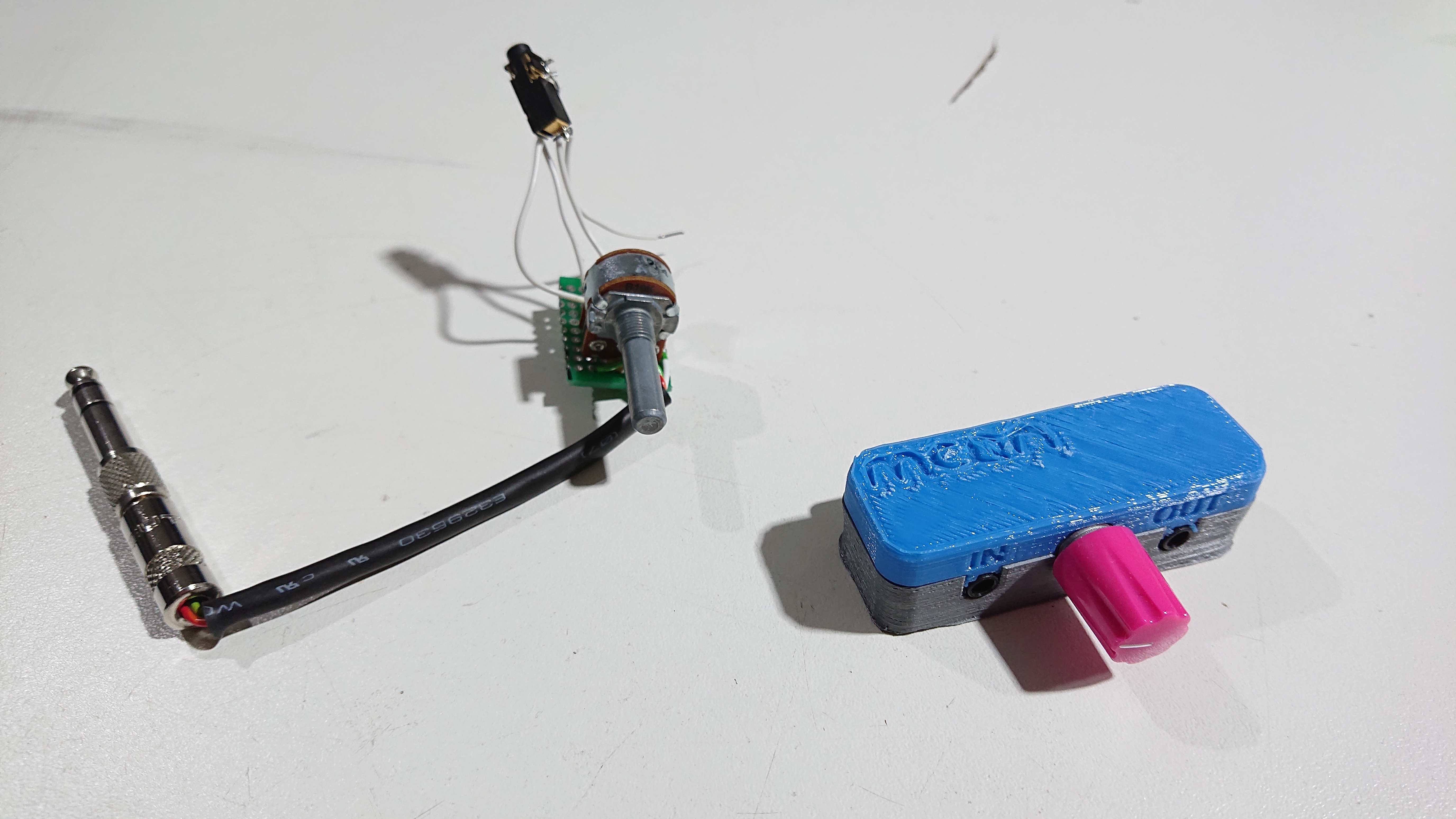 Two circuits on a table.  On the left is one with bare components fastened to a tiny rectangle of prototyping board, with a cable coming off to an audio jack plug and a 3.5mm jack socket suspended in mid-air on the wires connecting it to the prototyping board.  On the right is a two-tone 3D-printed plastic case with the MCQN logo on the blue top half and two 3.5mm audio sockets visible on the side of the case, labelled 'IN' and 'OUT'; between the sockets is a pink volume knob.