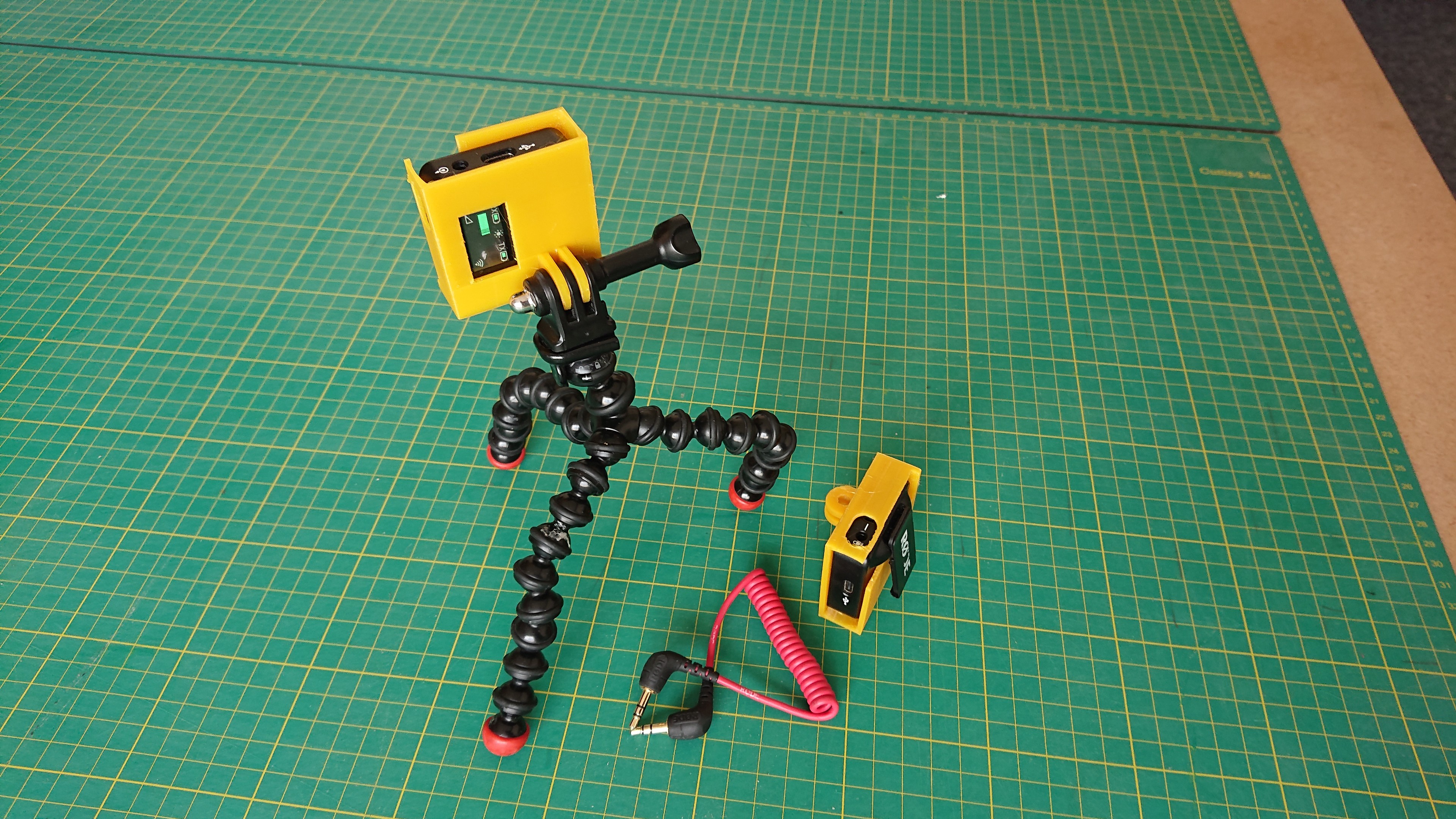 A small rectangular yellow box sits on top of a Gorillapod tripod, next to another one on a table but turned round. There's a hole in the top of them so a rectangular mic module can slide in, and a slot to allow the clip from the module to hold it in place.  At the base of the yellow boxes are the two protruding slots to mount on a GoPro mount; and there are cutouts allowing access to the power button, etc.