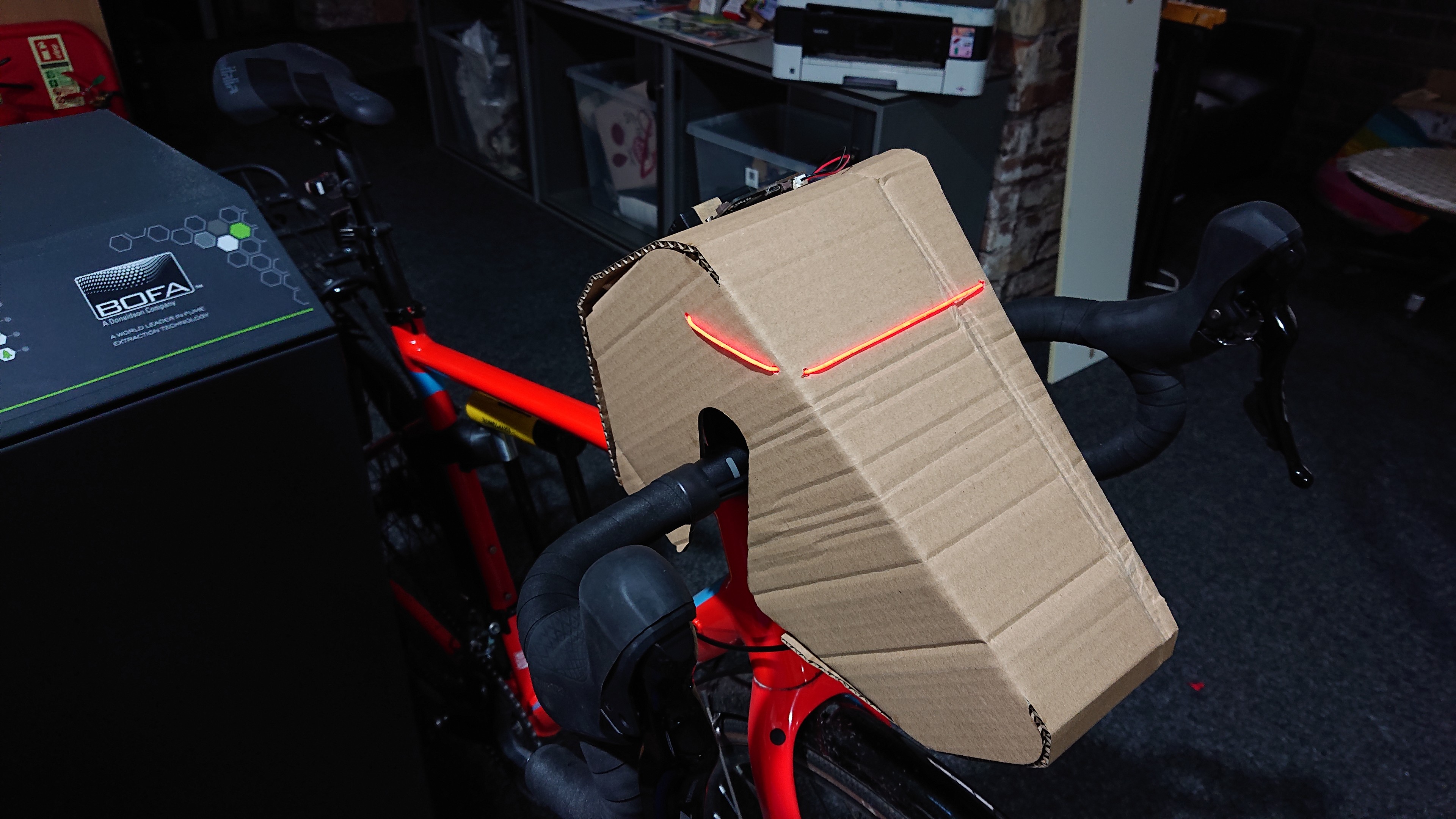 Close up of the laser-cut cardboard 'bike head' that looks a bit like K-9 from Doctor Who, mounted on the handlebars of a gravel bike.  There's a thin red illuminated strip running from half-way across the side and then all the way across the front, roughly where you'd imagine the eyes might be.