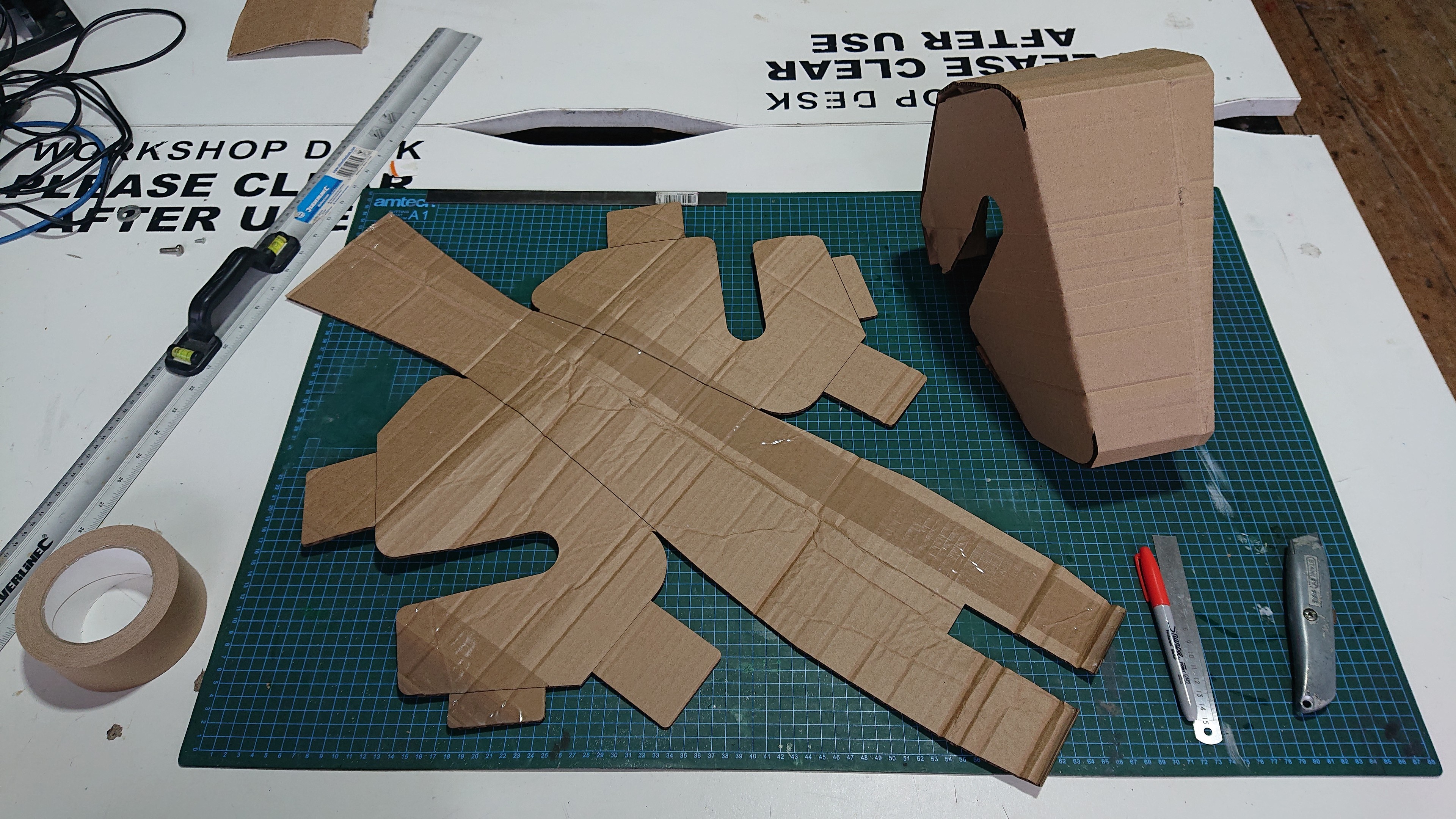 A workbench with a cutting mat, some tape, a Sharpie, a Stanley knife and a metal ruler.  On the cutting mat is a flat sheet of brown corrugated cardboard cut out in the shape of the unwrapped mesh from the CAD design.  Next to that is a version which has been folded up to make the triangular wedge shape.