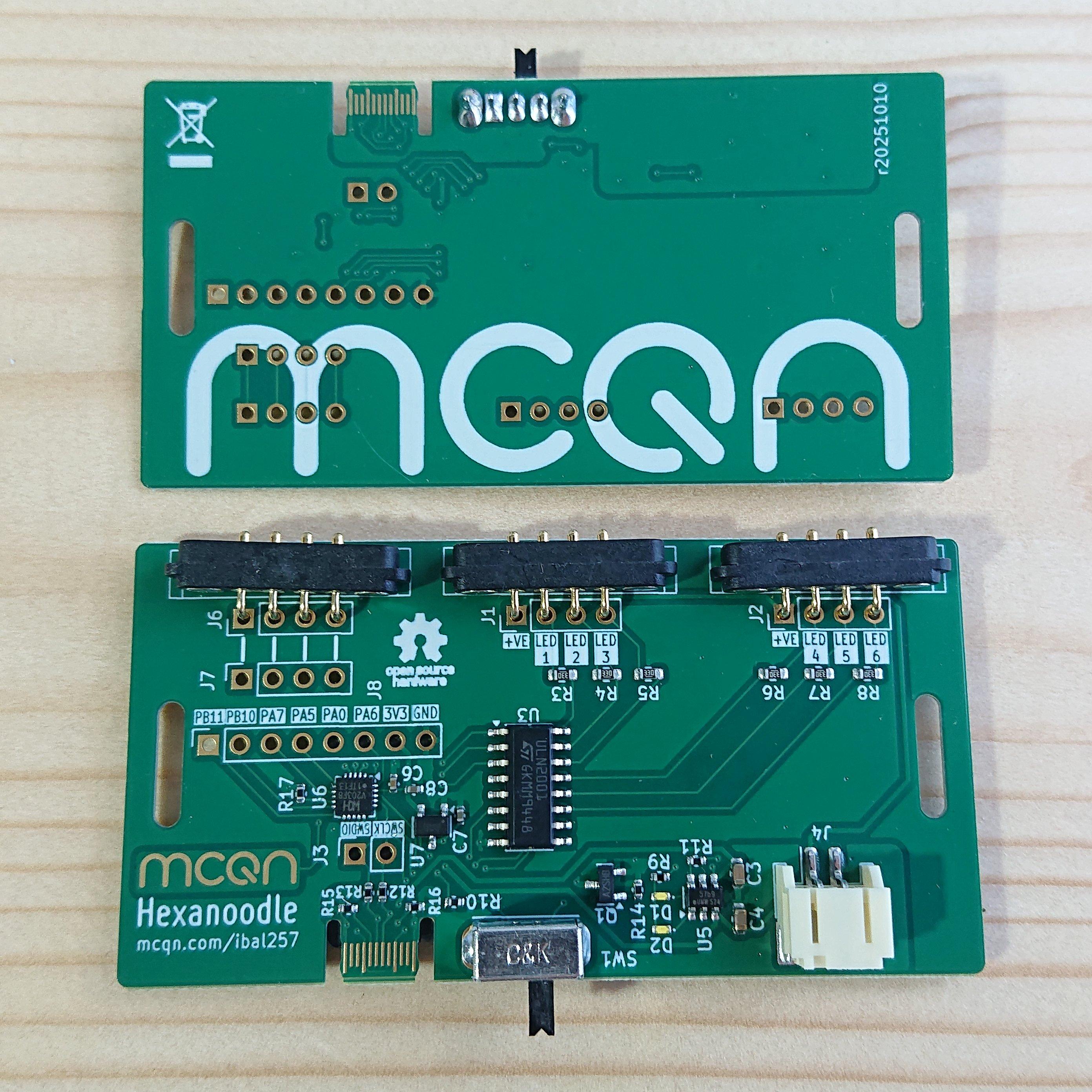Two rectangular printed circuit boards sat on a wooden table.  There's two so you can see both sides of the board: the underside is emblazoned with the MCQN logo; the top-side is where the action happens.  It has a few chips on it, a slide on/off switch, USB-C connector and rechargeable battery connector running along one side, and three 4-pin magnetic pogo-pin connectors running along the other side.  In the bottom left corner of the top-side is a gold MCQN logo (which will, hopefully, work as a capacitive touch pad) above the text Hexanoodle and mcqn.com/ibal257