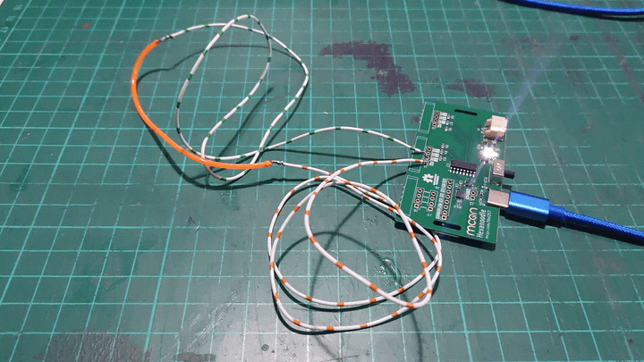 A green PCB sits on a cutting mat. A USB cable is plugged into it and a pair of wires come out of the other side of the board to an orange LED noodle.  A switch next to the USB cable gets switched on and the LED noodle lights up bright pink and flashes on and off at half-second intervals