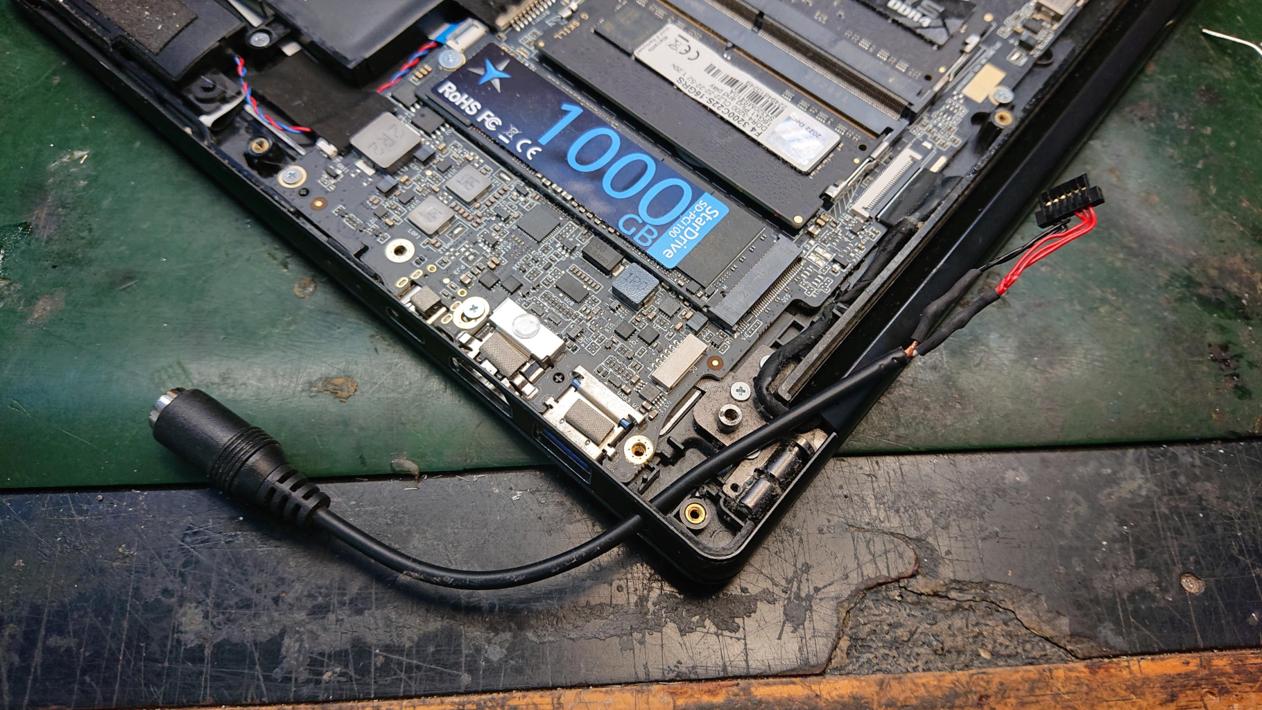 An opened laptop on a green mat. The image shows a repair to the charging connector, with a a black wire snaking across the circuit board, ready to be fitted. 