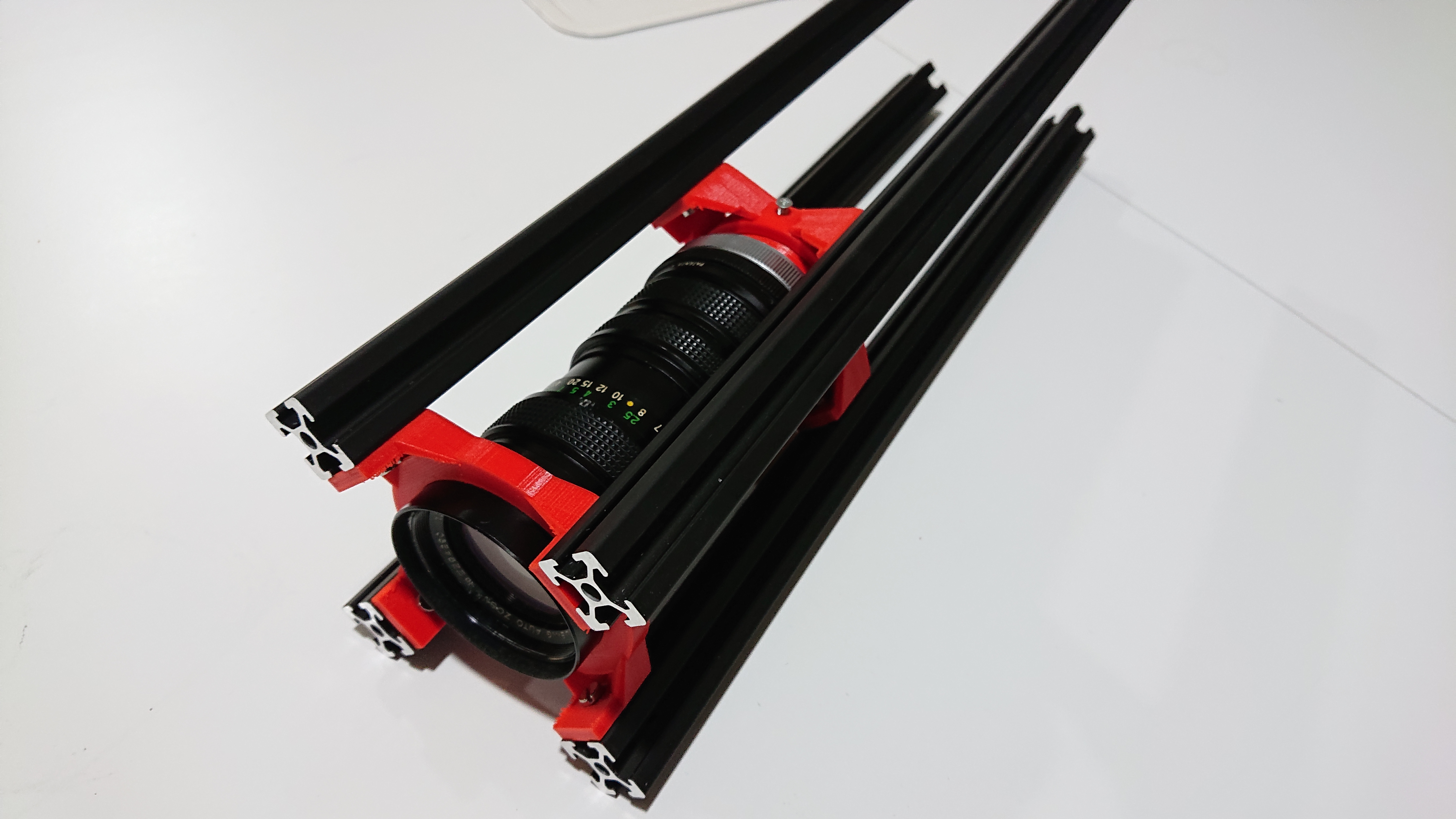 A partially-assembled ligra projector.  Four lengths of aluminium extrusion surround a couple of red 3D prints; the 3D prints hold a second-hand SLR telephoto lens.