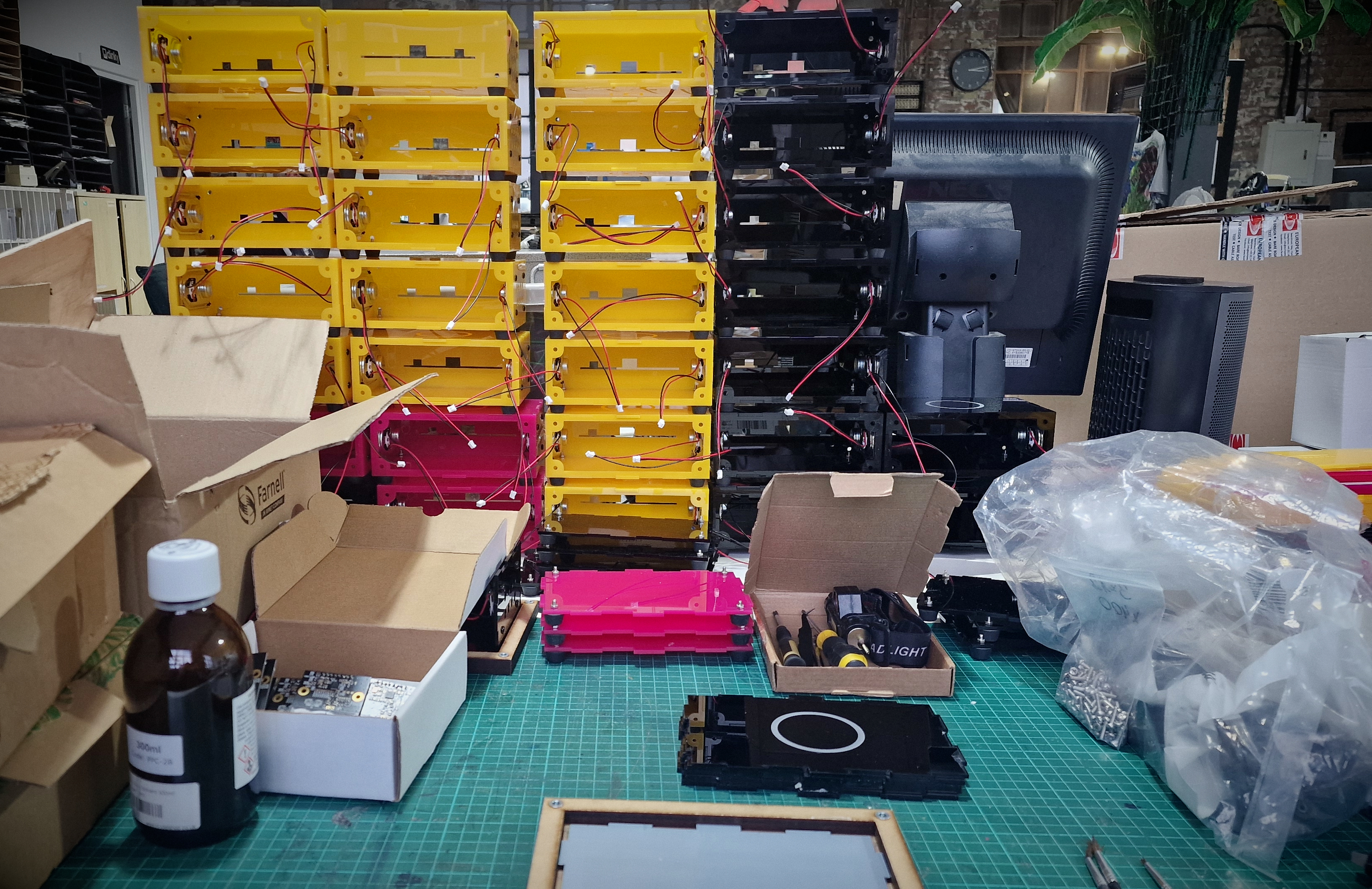A desk at DoES set up to assemble Museum in a Box boxes. In the background is a wall of yellow, pink and black cases with speaker wires hanging out. In the foreground on a green cutting mat are the tools needed to assemble them.