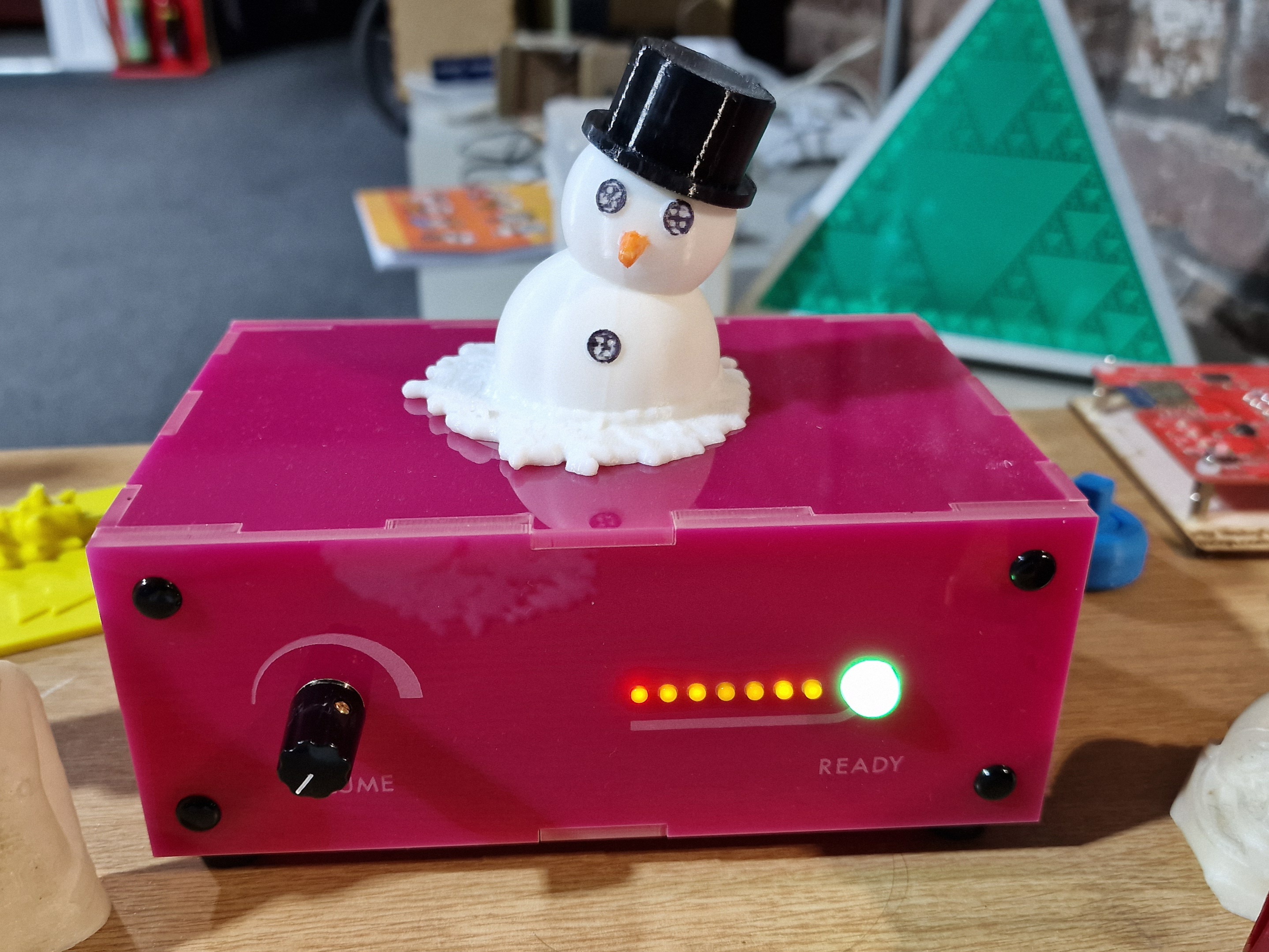 A 3d print of a partially melted snowman wearing a black top hat, sitting on a pink Museum in a box