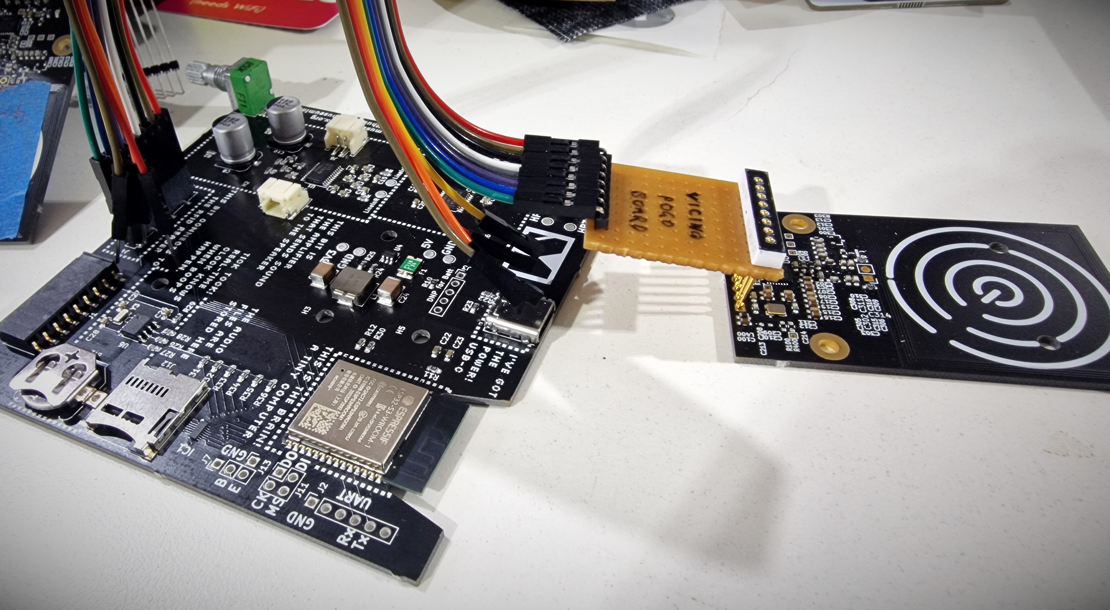 A black museum in box mainboard connected to a rainbow coloured ribbon of cable. This in turn is connected to some veroboard with pogo pins at the other end. These are being used to back a temporary connection to a Vicino board.