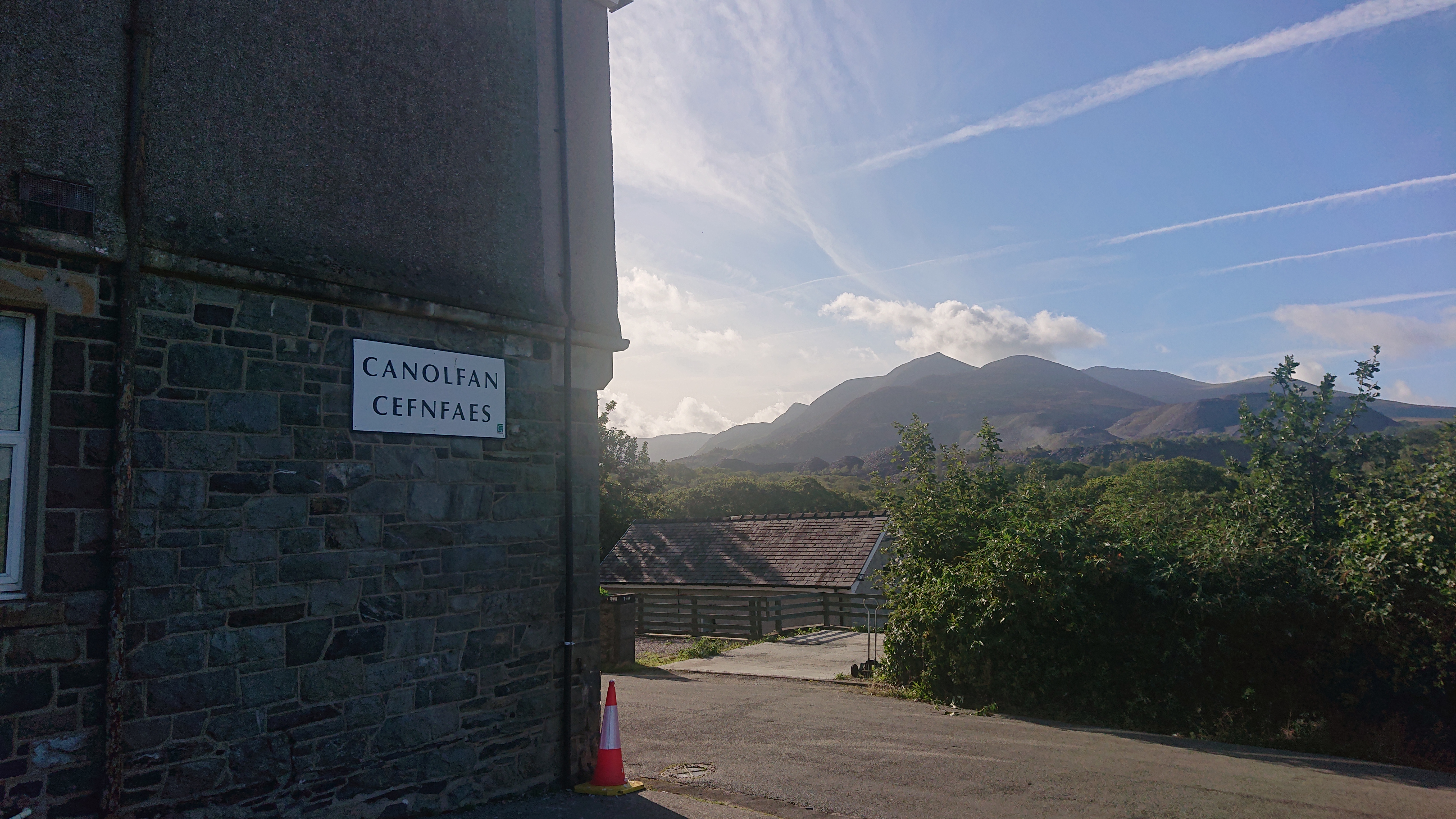Looking out across a Welsh valley, with a stone building to our left.  There's a sign on the building reading 'Canolfan Cefnfaes', its lower half is slate and on the top half the slate is hidden (I assume) behind render.  There's a bright blue sky with some contrails and bits of cloud here and there, and in the distance a range of beautiful mountain peaks.