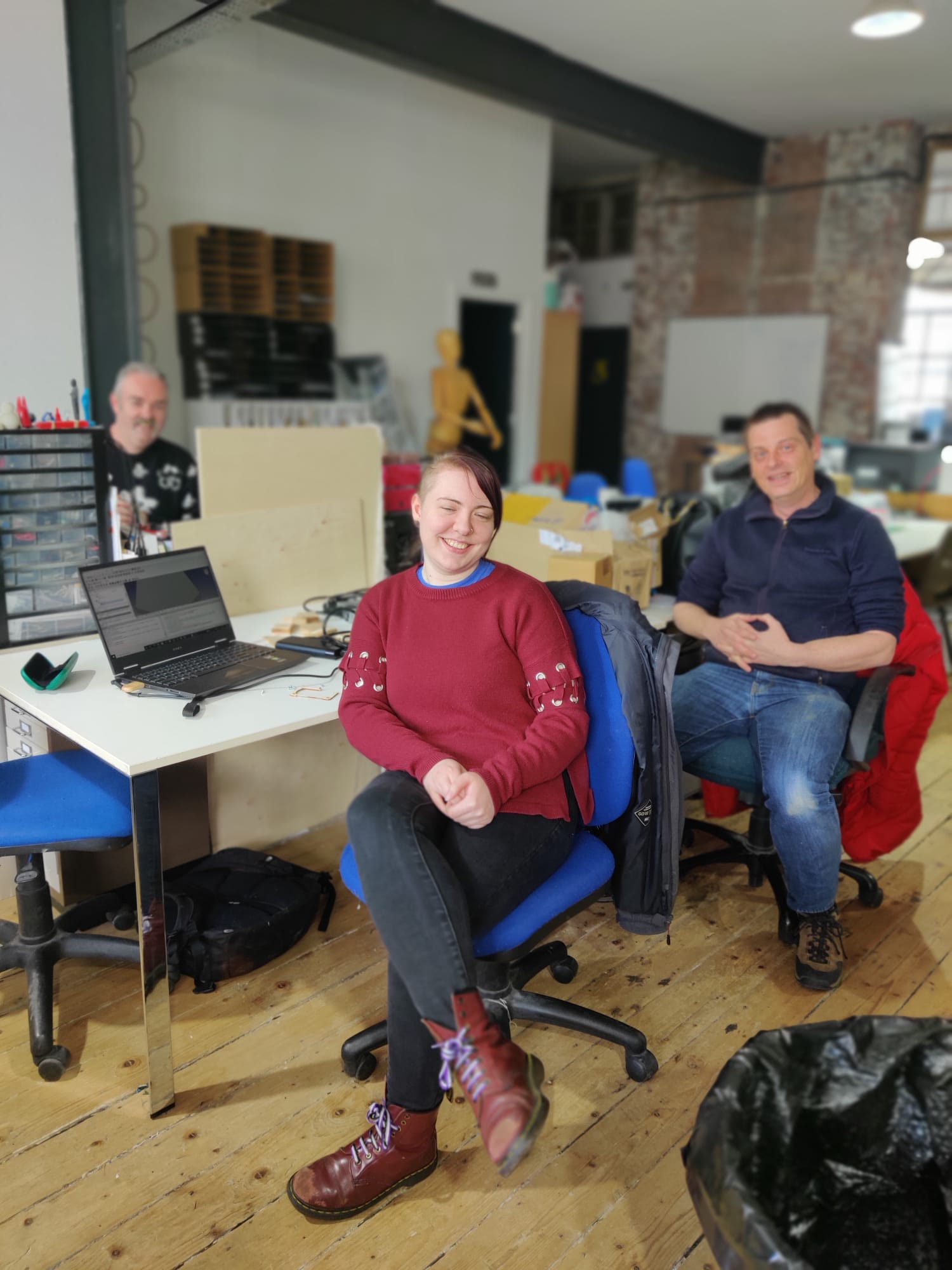 A view of an office, with wooden floorboards and white desks littered with laptops and other equipment.  There are three people visible, sat at their desks and facing the camera; a woman is centre of the frame and nearest the camera, flanked by two men, one on each side.