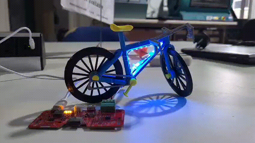 A small 3D-printed bike is sat on a desk next to a red circuit board. There's a tiny strip of RGB LEDs running round the inner triangle of the bike's frame, which are cycling through a rainbow of colours. The camera pans back to reveal a banana, which is larger than the whole bike.